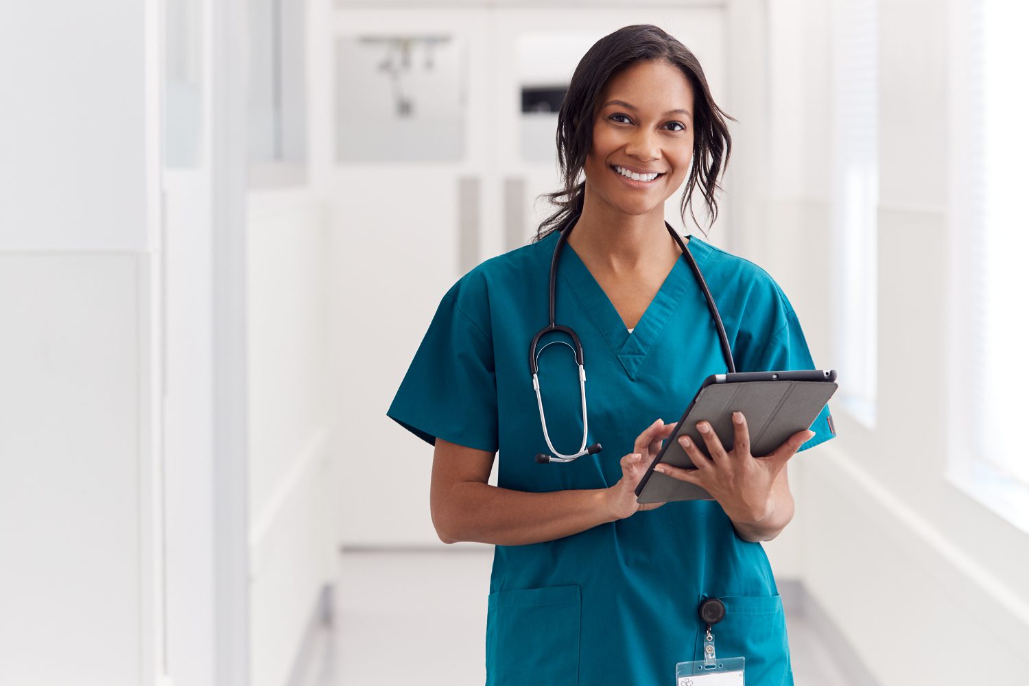 Portrait Of Smiling Female Doctor Wearing Scrubs In Hospital Corridor Holding Digital Tablet