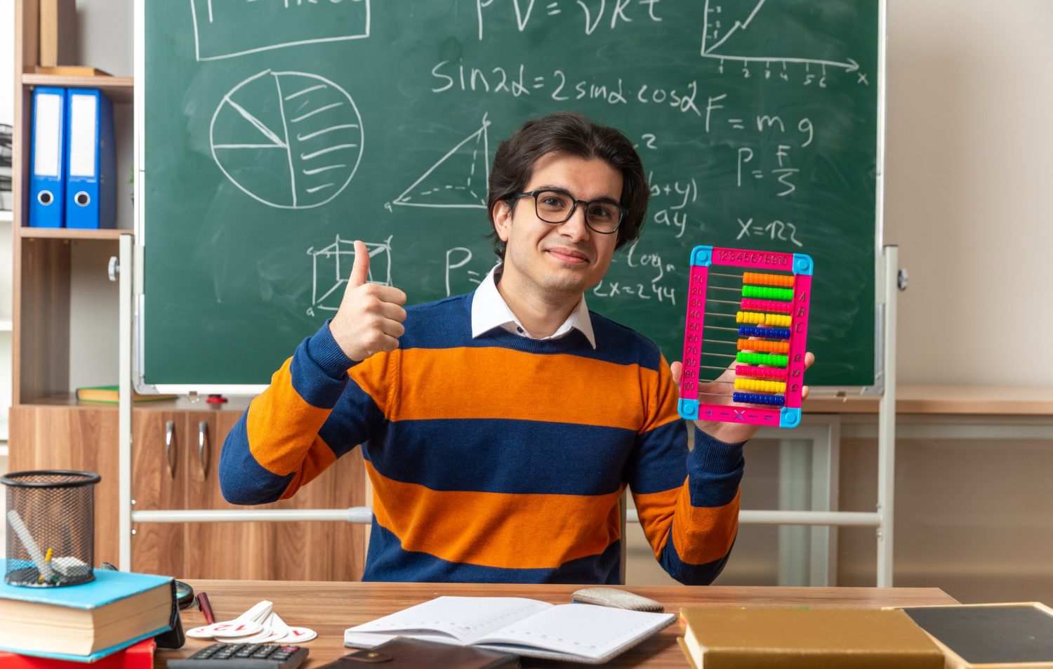 pleased young caucasian geometry teacher wearing glasses sitting at desk with school tools in classroom showing abacus and thumb up looking at camera